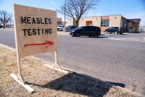 Outdoor sign directing people to a measles testing site, with vehicles and a clinic building in the background.