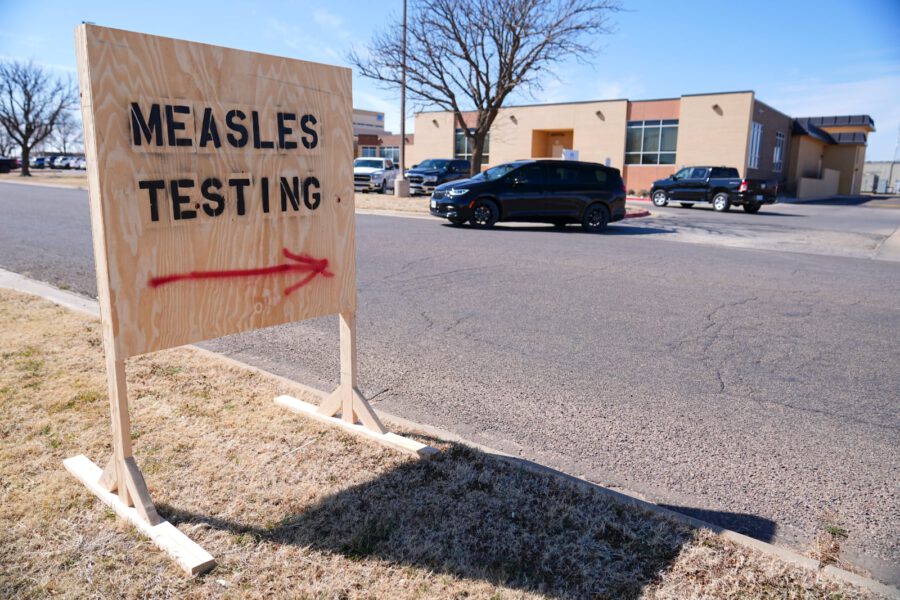 Outdoor sign directing people to a measles testing site, with vehicles and a clinic building in the background.