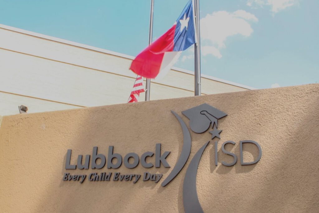 Lubbock ISD building sign with Texas and American flags flying in the background under a blue sky.