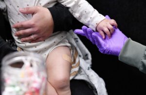 A child holds hands with a gloved healthcare worker after receiving a vaccination, indicated by a bandage on their thigh, during a measles outbreak in Texas amid rising vaccine exemptions and legislative changes affecting school immunization requirements.