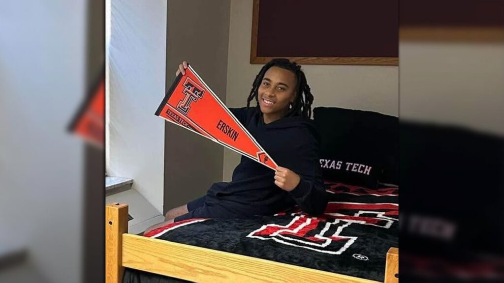 Young man smiling while sitting on a dorm bed with Texas Tech University bedding, holding a red and black pennant that reads “Erskin” and “Texas Tech.”