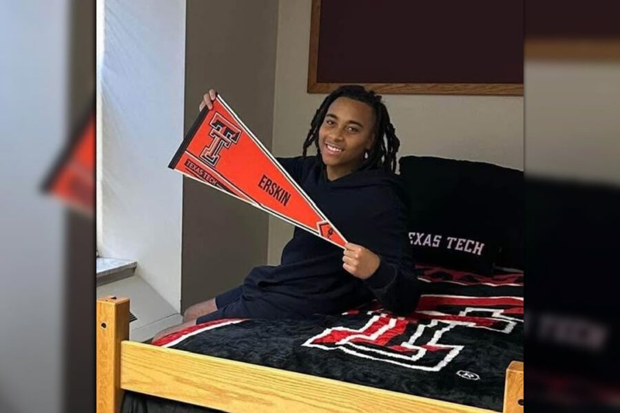 Young man smiling while sitting on a dorm bed with Texas Tech University bedding, holding a red and black pennant that reads “Erskin” and “Texas Tech.”
