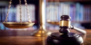 Close-up of a judge's gavel and brass scales of justice on a courtroom desk with law books in the background.
