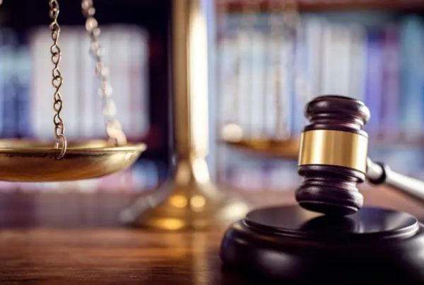 Close-up of a judge's gavel and brass scales of justice on a courtroom desk with law books in the background.