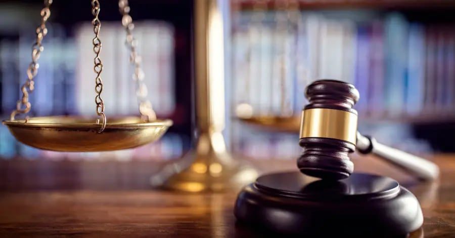 Close-up of a judge's gavel and brass scales of justice on a courtroom desk with law books in the background.
