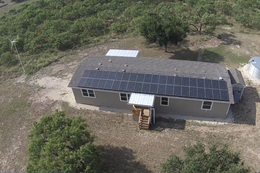 Aerial view of an off-grid home in Texas featuring rooftop solar panels, a wind turbine, and a rainwater collection tank as part of the HUMS (Home Utility Management System) sustainable energy and water initiative.
