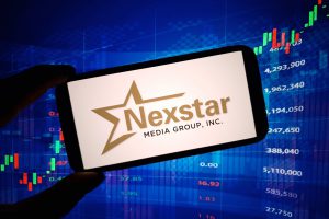 Nexstar Media Group logo on a smartphone screen with stock market charts in the background, symbolizing media consolidation and corporate growth.