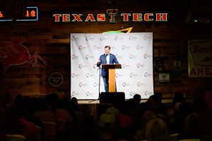 Brandon Creighton speaks at a Turning Point USA rally at Cook’s Garage in Lubbock, Texas, standing at a podium beneath the illuminated Texas Tech sign before a crowd.