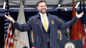 Vice President JD Vance speaking at a public event, wearing a navy jacket and yellow tie, gesturing with open arms in front of U.S. flags and a podium.
