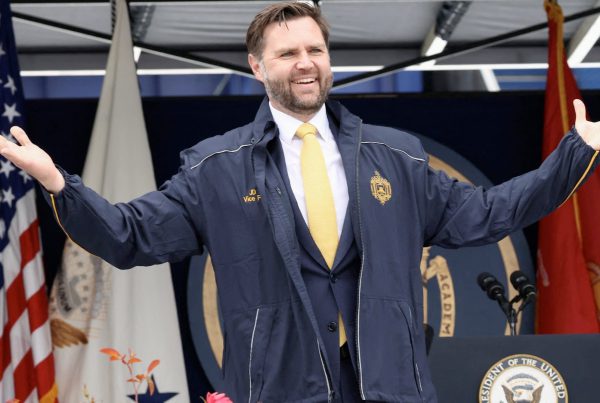 Vice President JD Vance speaking at a public event, wearing a navy jacket and yellow tie, gesturing with open arms in front of U.S. flags and a podium.