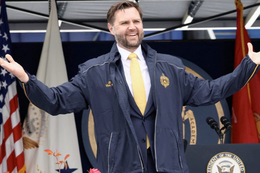 Vice President JD Vance speaking at a public event, wearing a navy jacket and yellow tie, gesturing with open arms in front of U.S. flags and a podium.