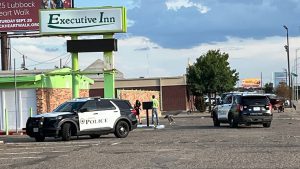 Two Lubbock Police SUVs parked outside the Executive Inn as officers speak with people near the boarded-up building under a bright green sign.