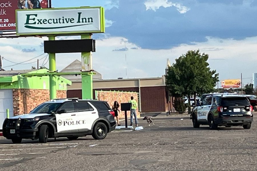 Two Lubbock Police SUVs parked outside the Executive Inn as officers speak with people near the boarded-up building under a bright green sign.