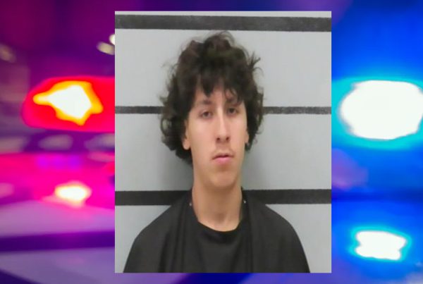Mugshot of Adrian Castilleja with curly hair in front of a height chart background, with police lights blurred behind him.