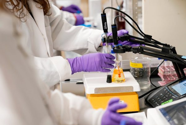 Scientists wearing lab coats and purple gloves testing water samples for PFAS contamination in a laboratory.