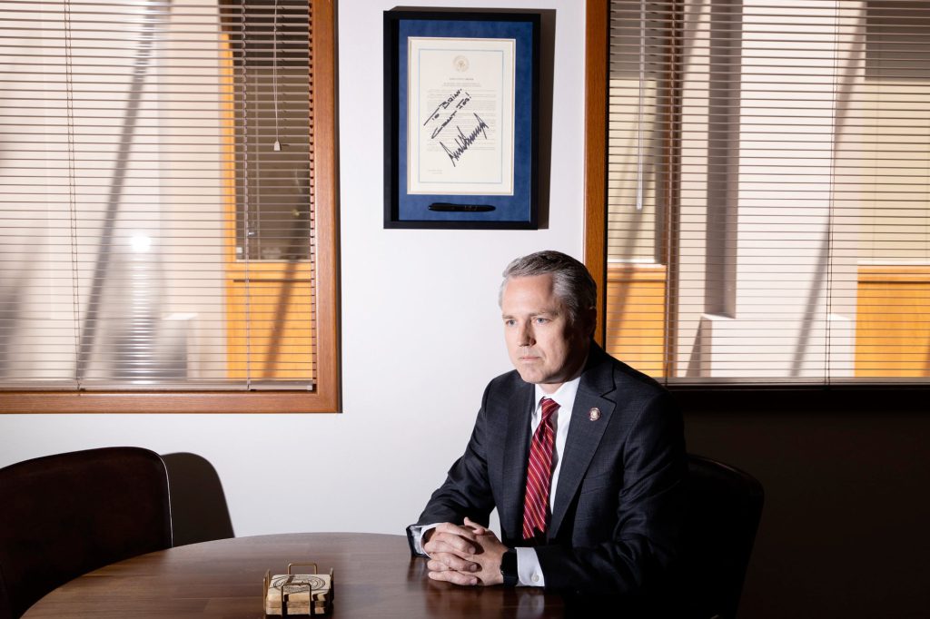 Texas Rep. Brian Harrison sits at a conference table in his office, hands clasped and expression stern, beneath a framed Trump-signed document and closed blinds casting sharp shadows across the room.
