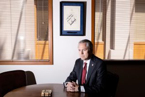 Texas Rep. Brian Harrison sits at a conference table in his office, hands clasped and expression stern, beneath a framed Trump-signed document and closed blinds casting sharp shadows across the room.