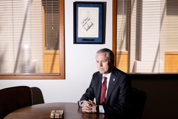 Texas Rep. Brian Harrison sits at a conference table in his office, hands clasped and expression stern, beneath a framed Trump-signed document and closed blinds casting sharp shadows across the room.