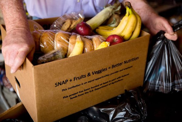 A person holding a cardboard box filled with groceries such as bananas, apples, corn, and bread labeled “SNAP + Fruits & Veggies = Better Nutrition,” symbolizing food assistance and nutrition support in Texas.