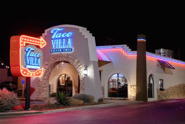 Exterior of Taco Villa in Lubbock, Texas at night, featuring neon red and blue lights on the stucco building and a glowing retro arrow sign.