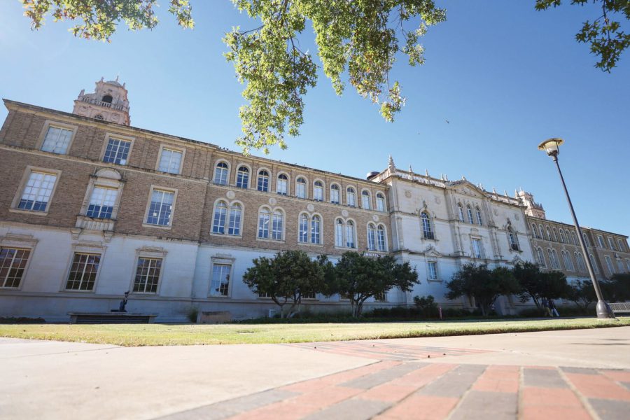 The Texas Tech University Administration Building in Lubbock, Texas, viewed from the campus lawn on a sunny day with clear blue skies and tree branches framing the scene.