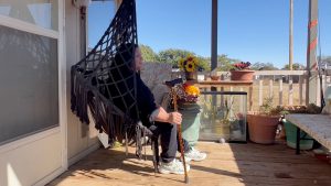 Ida Bosque sitting on her porch swing at a Lubbock mobile home, holding a cane beside potted plants and sunflowers on a sunny day.