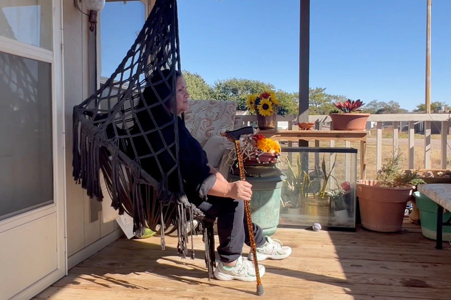 Ida Bosque sitting on her porch swing at a Lubbock mobile home, holding a cane beside potted plants and sunflowers on a sunny day.