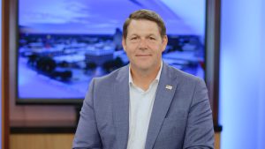 Jodey Arrington seated in a Lubbock TV studio wearing a blue blazer with an American flag lapel pin, smiling in front of a blurred newsroom backdrop.