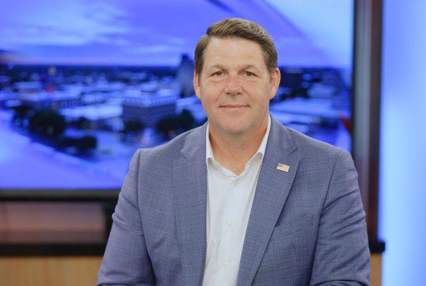 Jodey Arrington seated in a Lubbock TV studio wearing a blue blazer with an American flag lapel pin, smiling in front of a blurred newsroom backdrop.