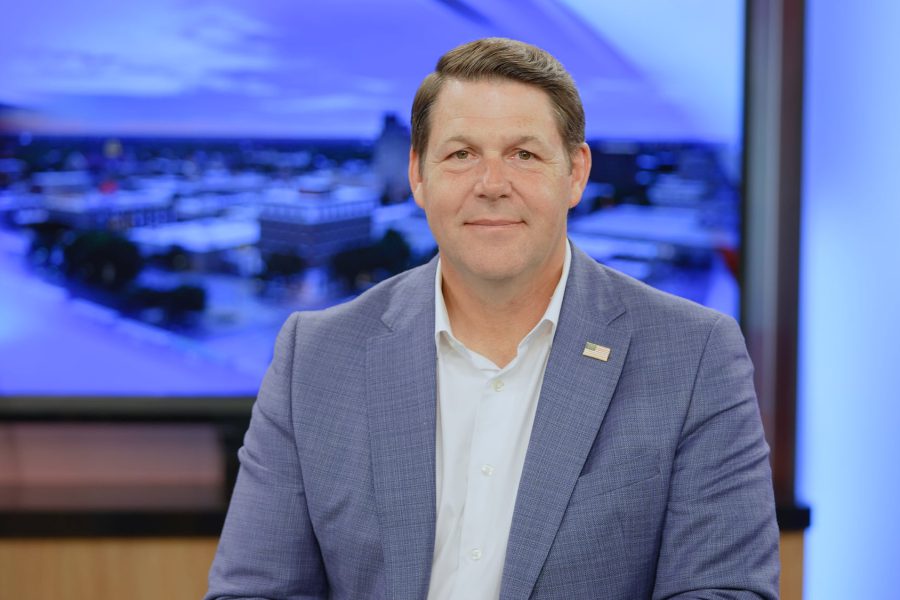 Jodey Arrington seated in a Lubbock TV studio wearing a blue blazer with an American flag lapel pin, smiling in front of a blurred newsroom backdrop.