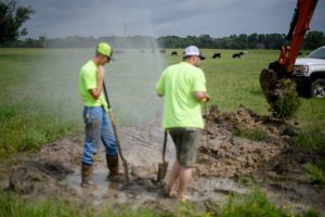 Two workers repair a leaking water line in a muddy rural Texas field as cattle graze in the background.