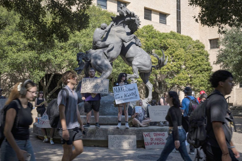 Students hold signs calling for free speech and administrative accountability during a campus protest in front of a large horse statue at a Texas university.