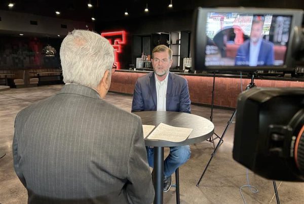 Two men seated at a small table during a televised interview at Texas Tech, with cameras recording and the iconic red Double T glowing in the background.