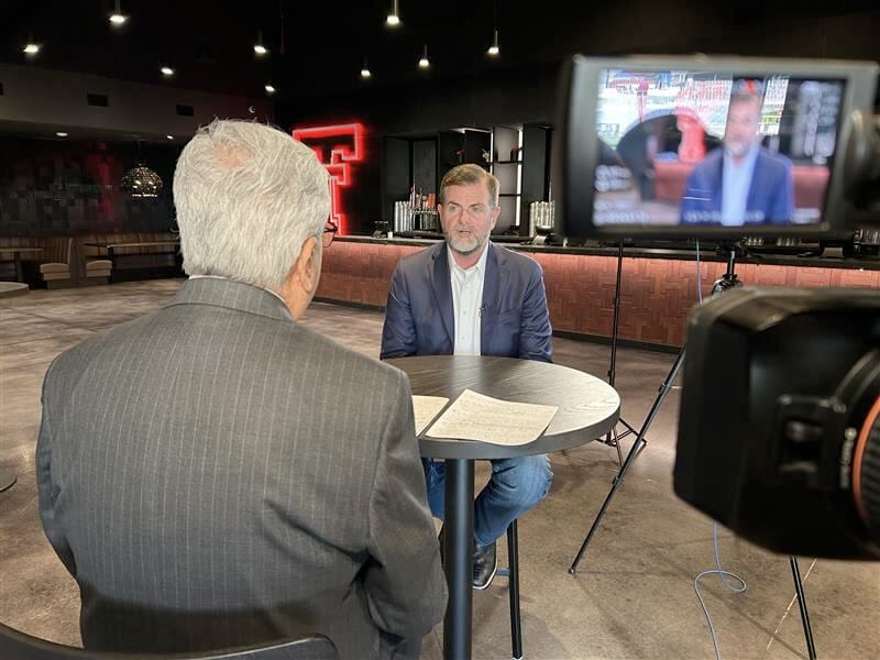 Two men seated at a small table during a televised interview at Texas Tech, with cameras recording and the iconic red Double T glowing in the background.