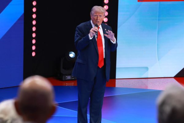 Donald Trump in a blue suit and red tie speaking onstage with a microphone during a televised event.