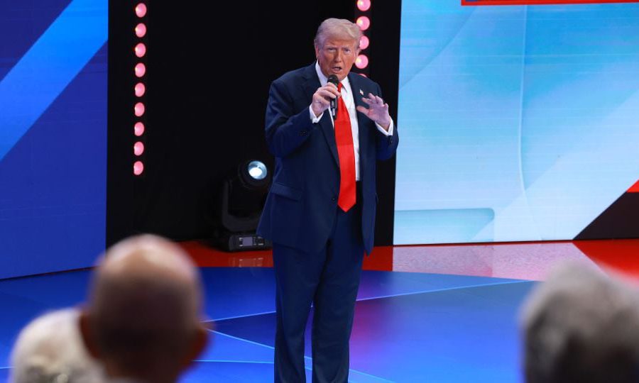 Donald Trump in a blue suit and red tie speaking onstage with a microphone during a televised event.