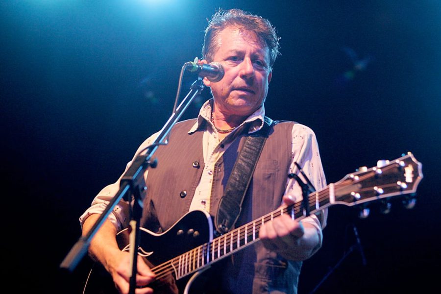 West Texas singer-songwriter Joe Ely performs live on stage, playing an acoustic guitar under blue stage lights.