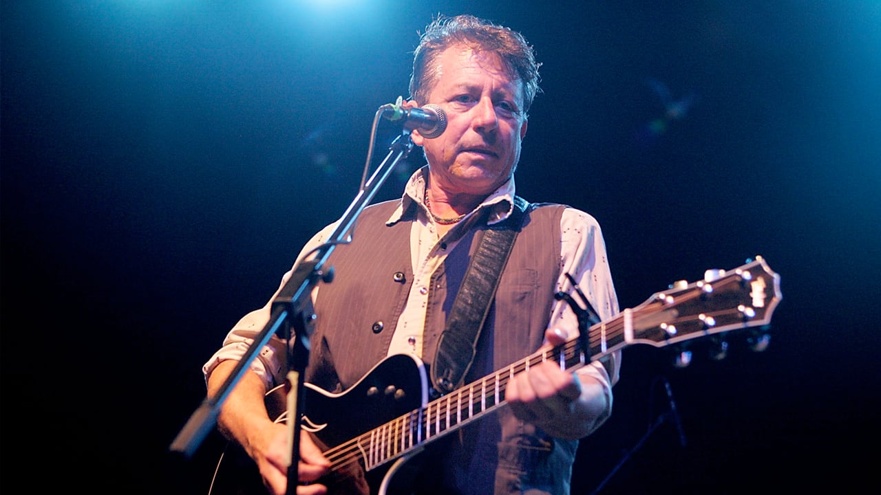 West Texas singer-songwriter Joe Ely performs live on stage, playing an acoustic guitar under blue stage lights.