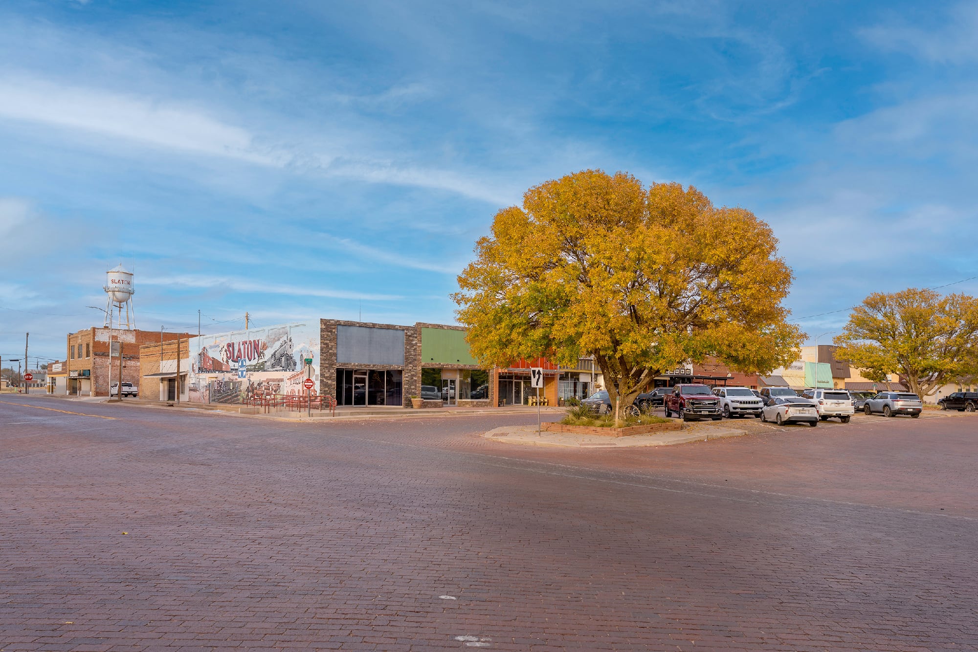 Downtown Slaton, Texas town square with brick streets, storefronts, fall trees, and a mural under a wide West Texas sky.