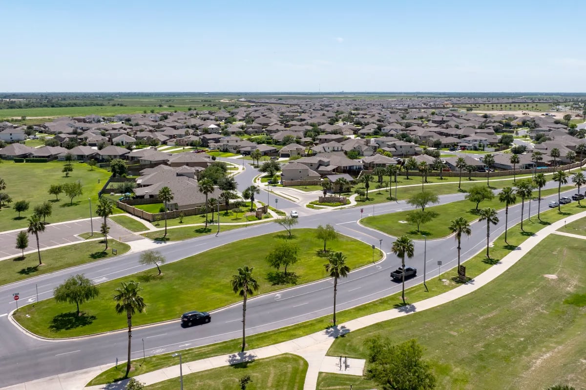 Aerial view of a sprawling South Texas suburban neighborhood with newly built homes, wide roads, palm trees, and undeveloped land stretching into the distance.