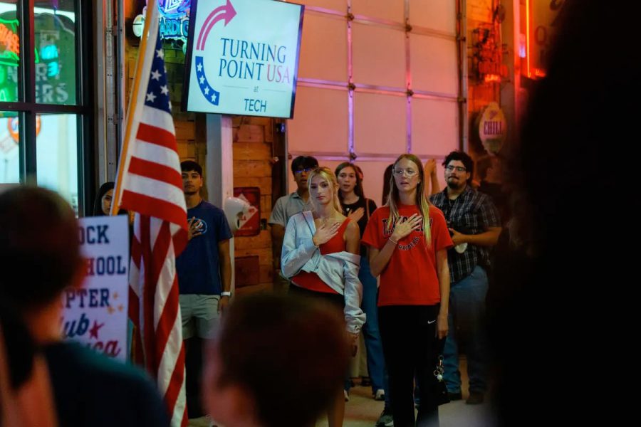Students stand with their hands over their hearts during a Turning Point USA gathering in Lubbock, with an American flag and TPUSA sign displayed in the background.
