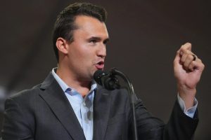 Conservative political activist Charlie Kirk speaking into a microphone on stage with a raised fist gesture.