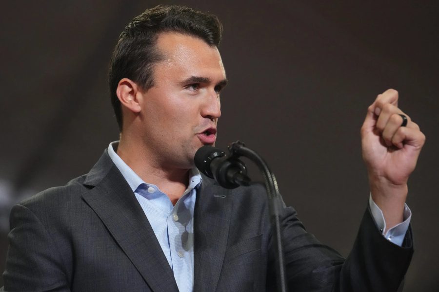 Conservative political activist Charlie Kirk speaking into a microphone on stage with a raised fist gesture.