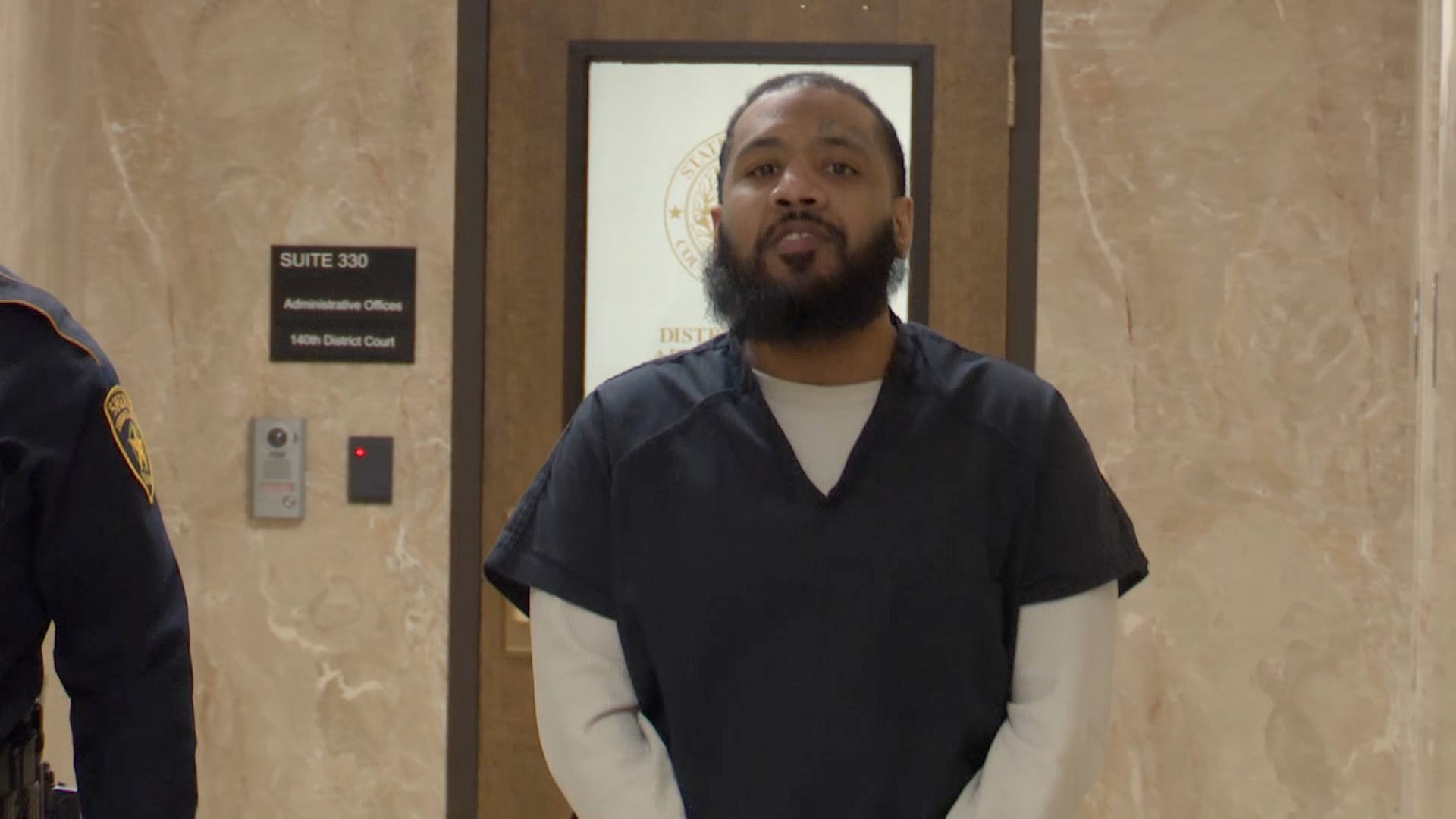 Henry Mena in a dark jail uniform standing in a courthouse hallway outside a district court office, with a law enforcement officer partially visible beside him.