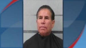 A booking photo of 70-year-old Raymundo Almaraz, wearing a black garment against a grey background with horizontal lines.