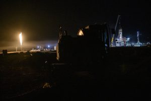 Nighttime oil and gas drilling site in West Texas with active flaring illuminating rigs and industrial equipment under a dark sky.