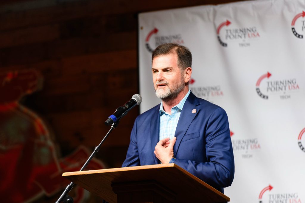 Texas Tech Chancellor Brandon Creighton, wearing a blue blazer and light blue checkered shirt, speaks at a podium during a Turning Point USA event. A white backdrop with the Turning Point USA logo is visible behind him.