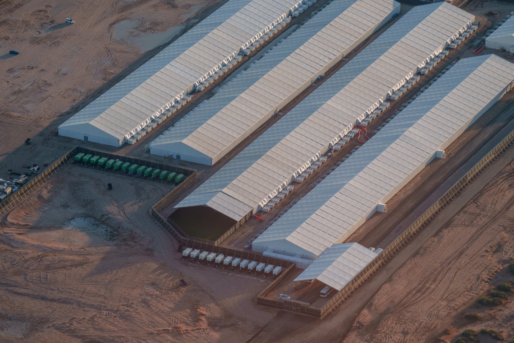 An aerial view of Camp East Montana at Fort Bliss, showing a massive complex of long, white industrial tents surrounded by dirt and security fencing. This "hastily built" 5,000-bed facility in El Paso is currently housing over 3,000 detainees amidst reports of medical neglect, tuberculosis outbreaks, and sewage issues.