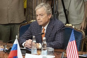Don Huffines sitting at a polished wooden table between a Russian flag and a US flag, looking remarkably like a man who just realized he's being photographed at the exact place his "America First" base would find most confusing.