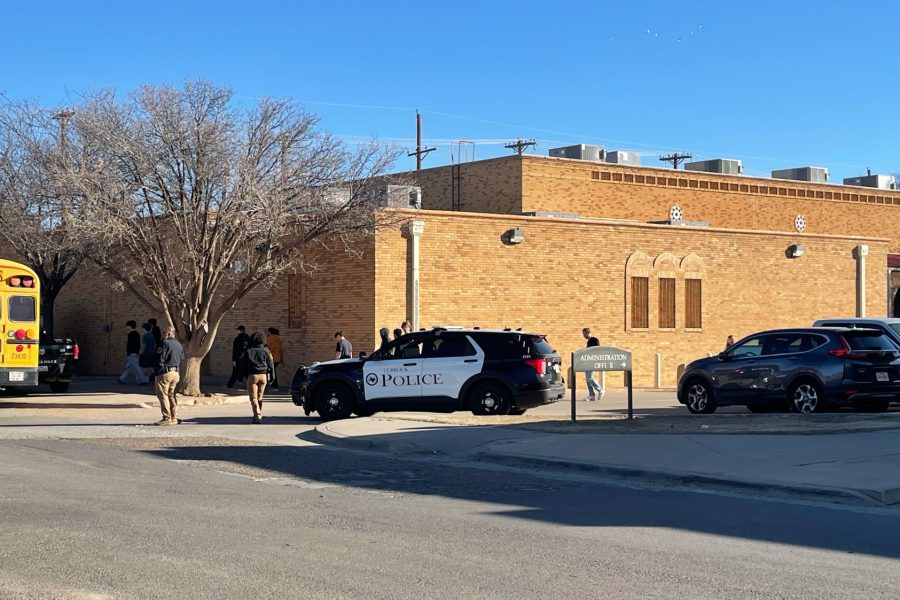 A Lubbock Police patrol car sits parked in front of Lubbock High School during a lockdown evacuation. Students and staff are seen walking toward a waiting yellow school bus while officers monitor the campus perimeter.